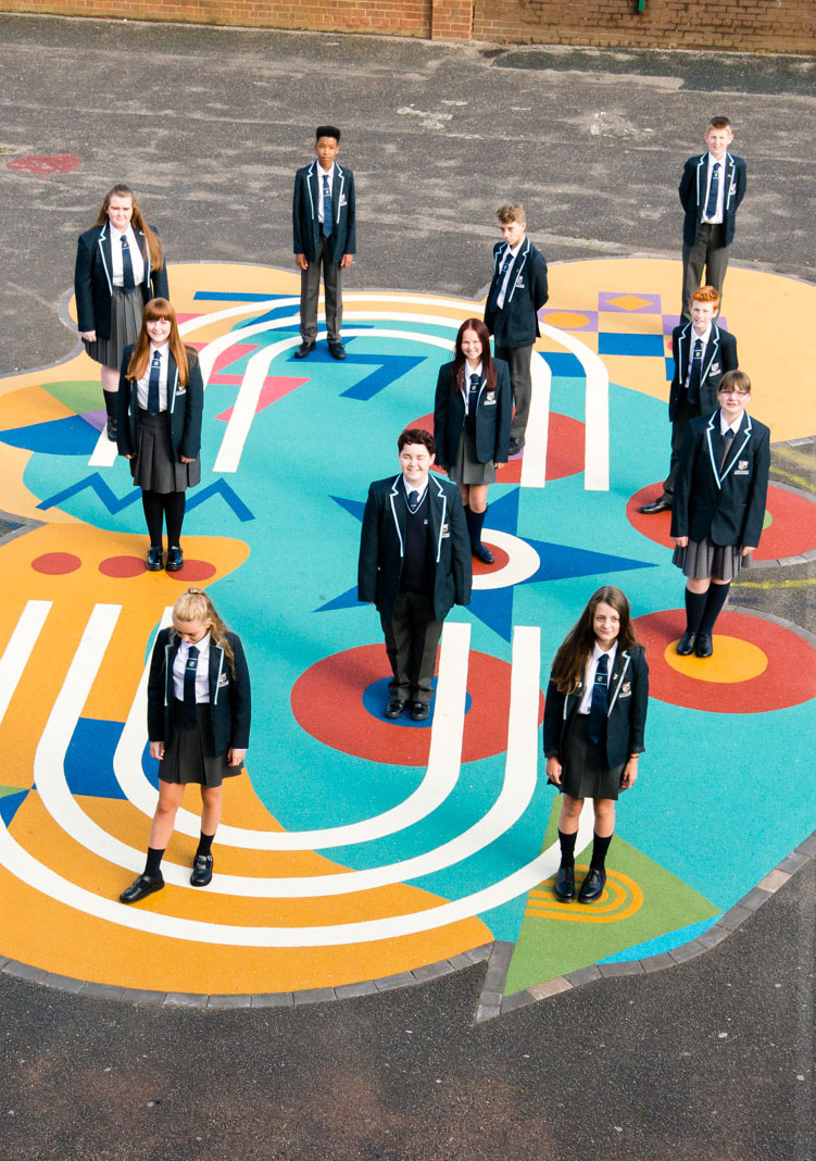 11 children smiling at the camera and stood on a piece of flamboyant art on the playground.