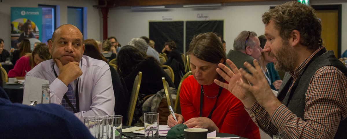 Three people sat in a busy conference room. They look to be debating a personal topic and making notes.