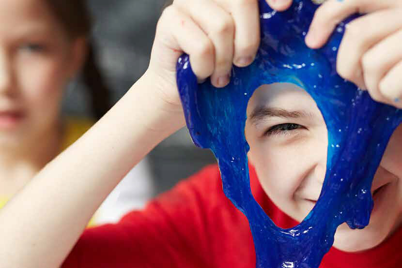 Young boy holding drooping blue playdough in front of his face. He's smiling and one of his eyes is peering through the playdough.