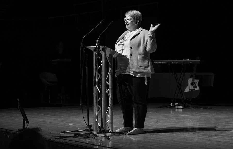 Black and white photo of a smartly dressed woman smiling and presenting on stage.