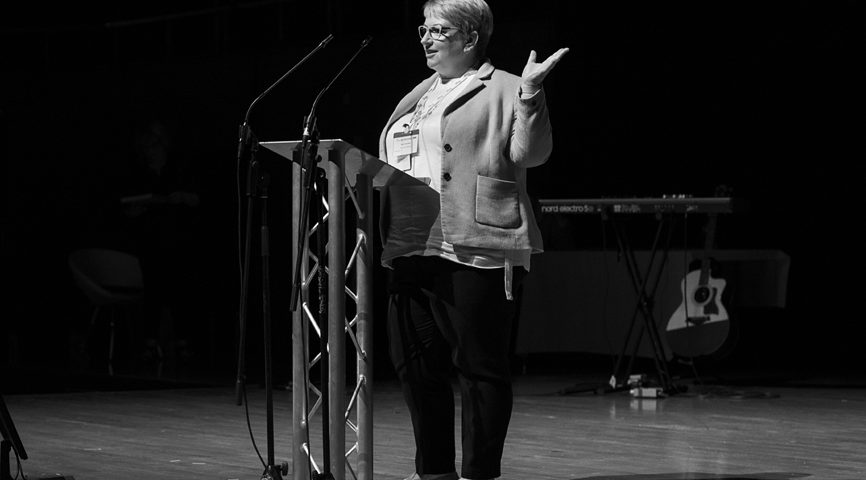 Black and white photo of a smartly dressed woman smiling and presenting on stage.