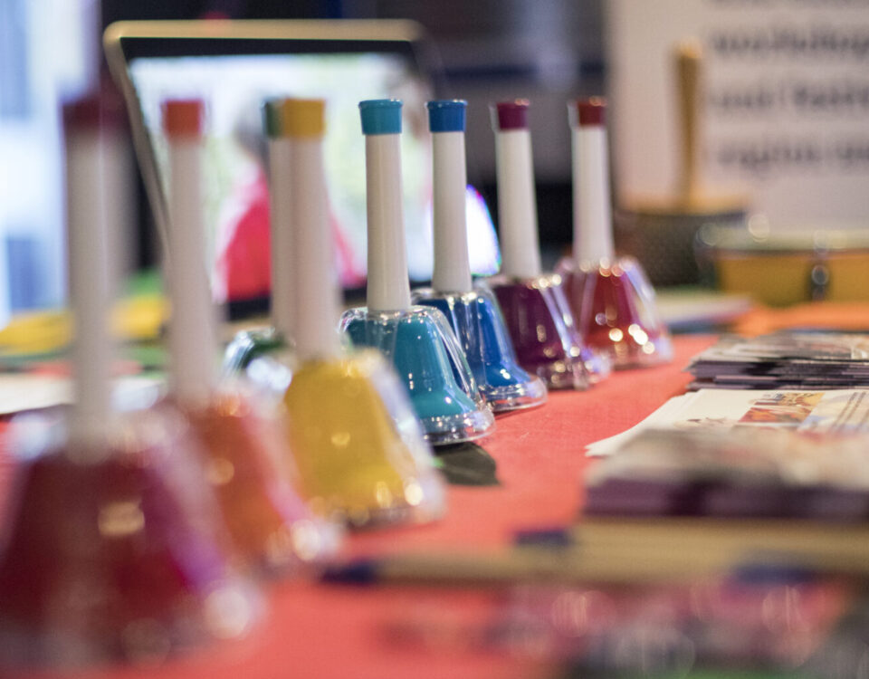 A row of colourful bells laid across a red table with pieces of paper out of focus on the end fo the table.