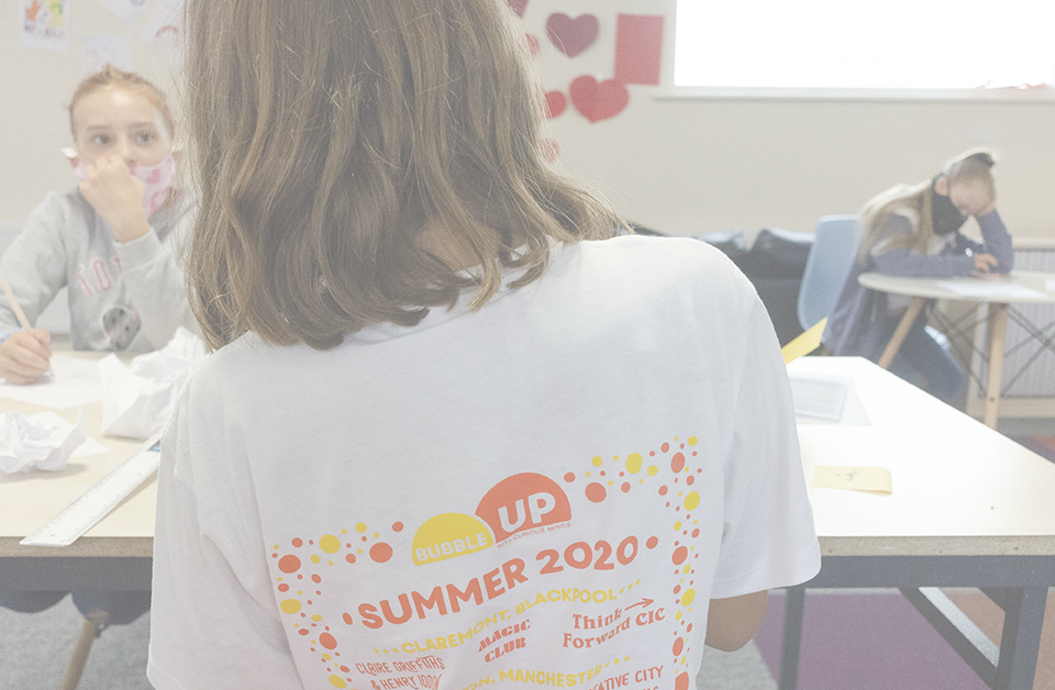 A young Bubble Up participant works at a table with a friend. Both are wearing face coverings. One is wearing a Bubble Up t-shirt.