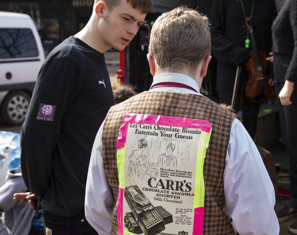 A young adult and older adult are in discussion. The older adult has an historic advert for biscuits taped to his back.