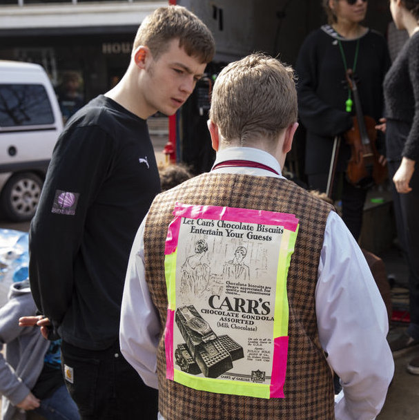 A young adult and older adult are in discussion. The older adult has an historic advert for biscuits taped to his back.