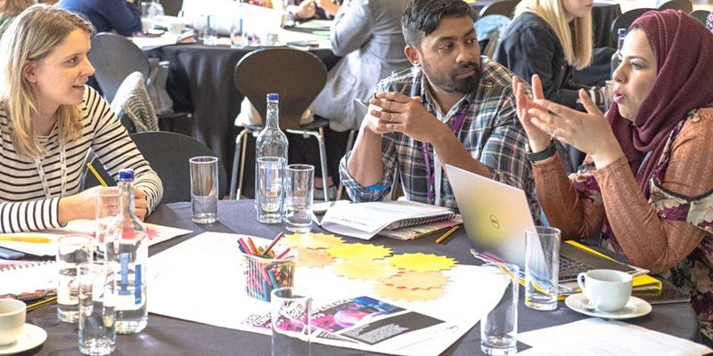 Three people are sat around a table in the midst of dicussion. They have a laptop and paper on the table in front of them.