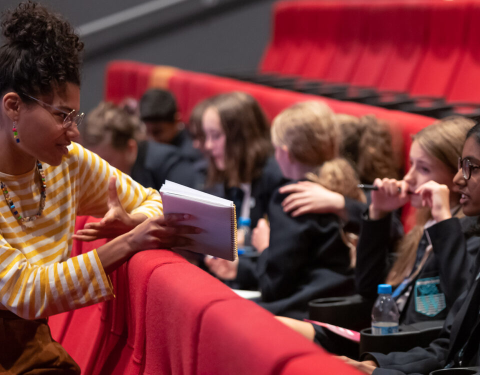 A teacher is with pupils in theatre seating. She is showing them a book and explaining something.