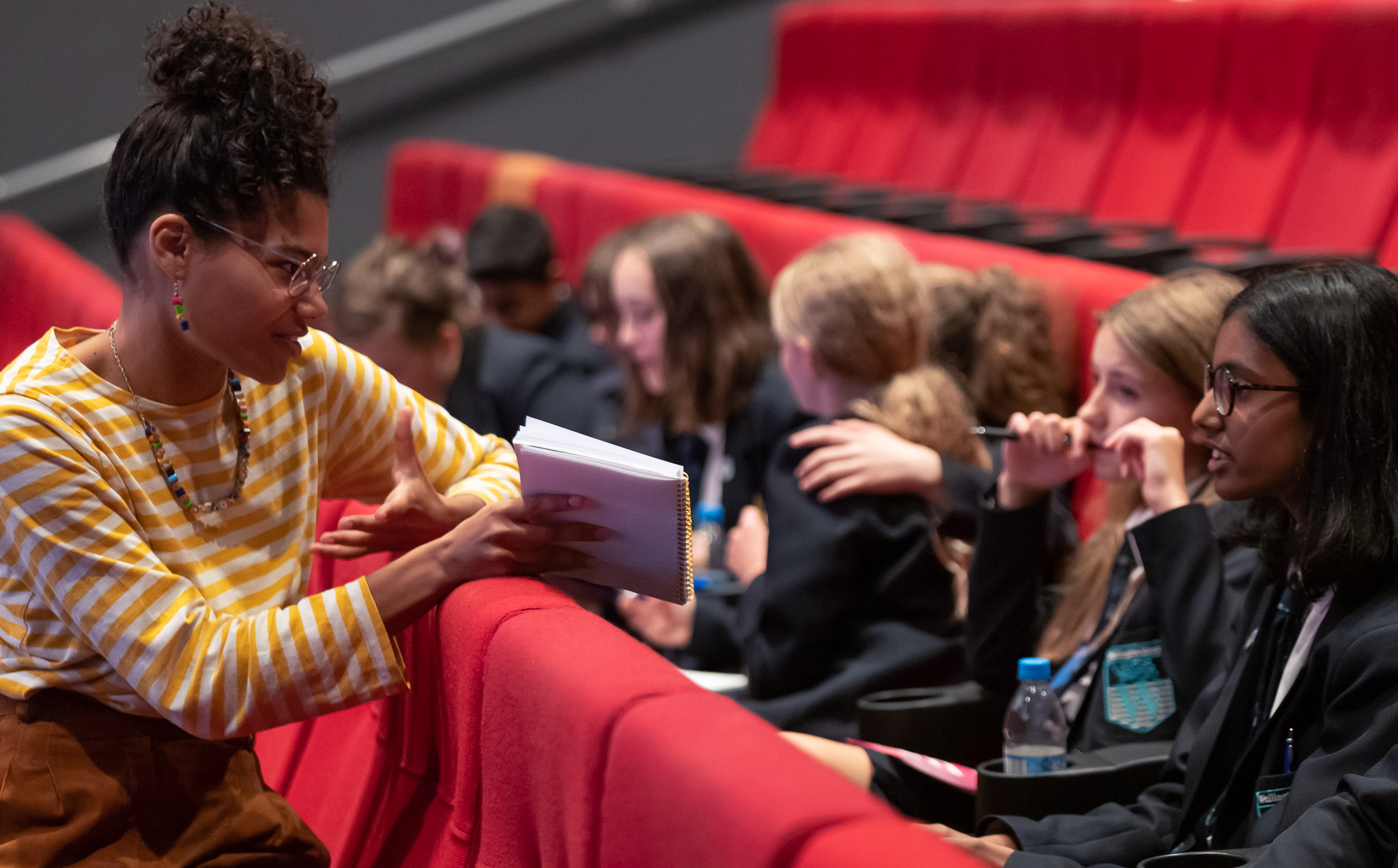 A teacher is with pupils in theatre seating. She is showing them a book and explaining something.