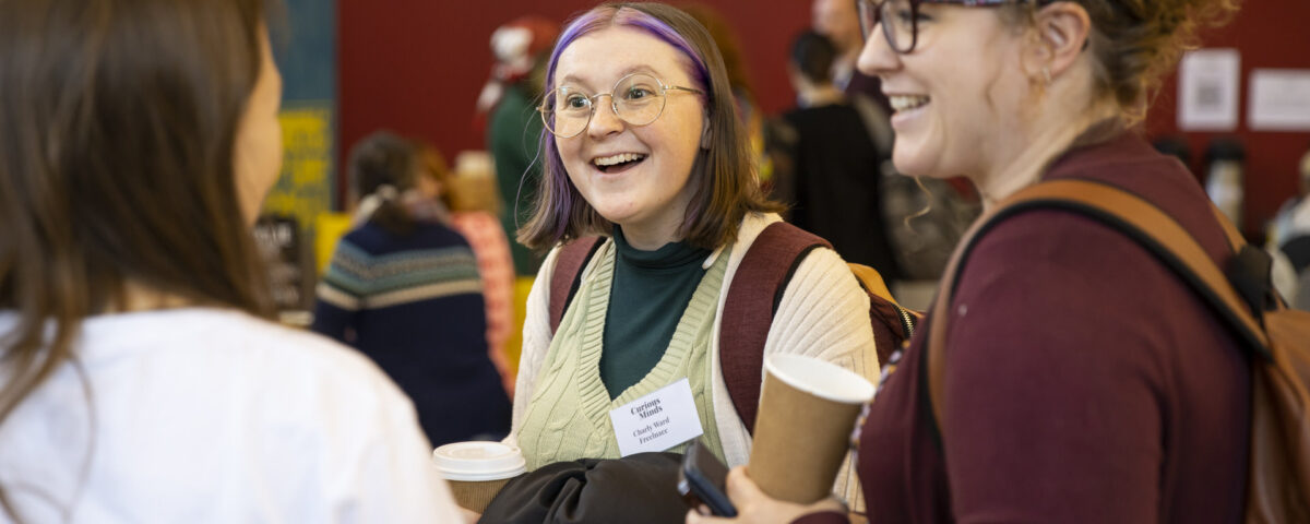 Holly Ball is chatting animatedly with two people at an event. She is wearing a burgundy top and carrying a coffee cup.