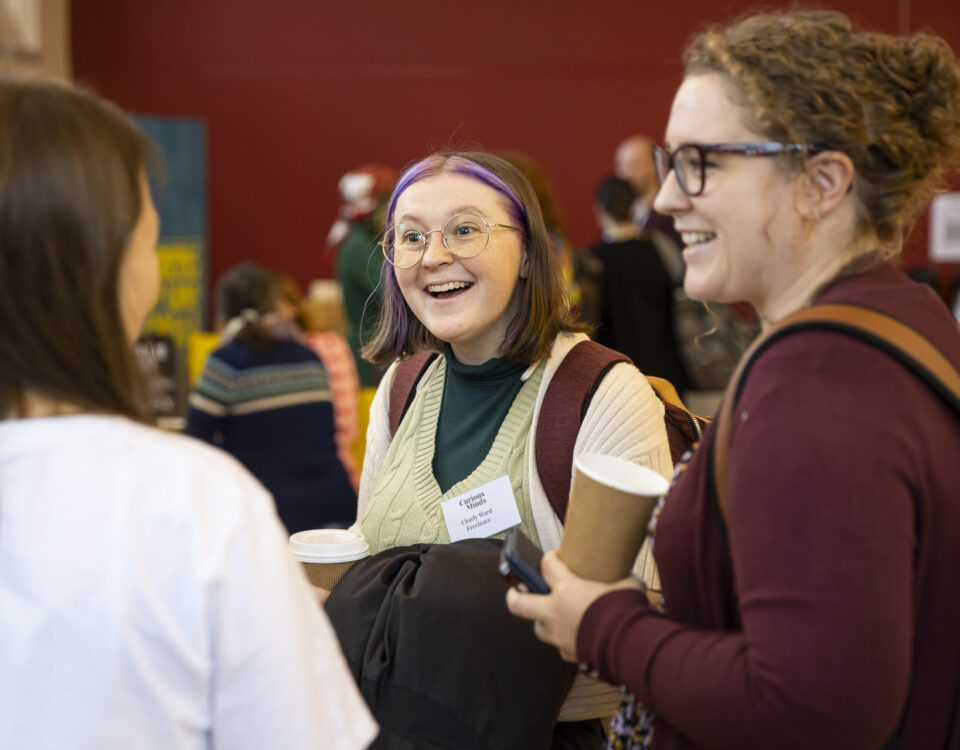 Holly Ball is chatting animatedly with two people at an event. She is wearing a burgundy top and carrying a coffee cup.