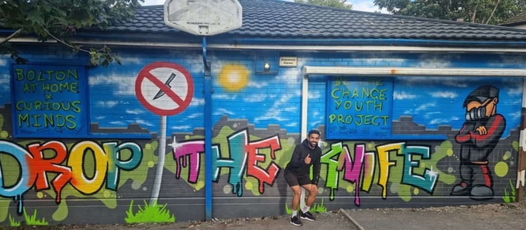 Image is a closeup of the Bolton@Home grafitti artwork. Faisal is stood in front looking delighted and giving the thumbs up.