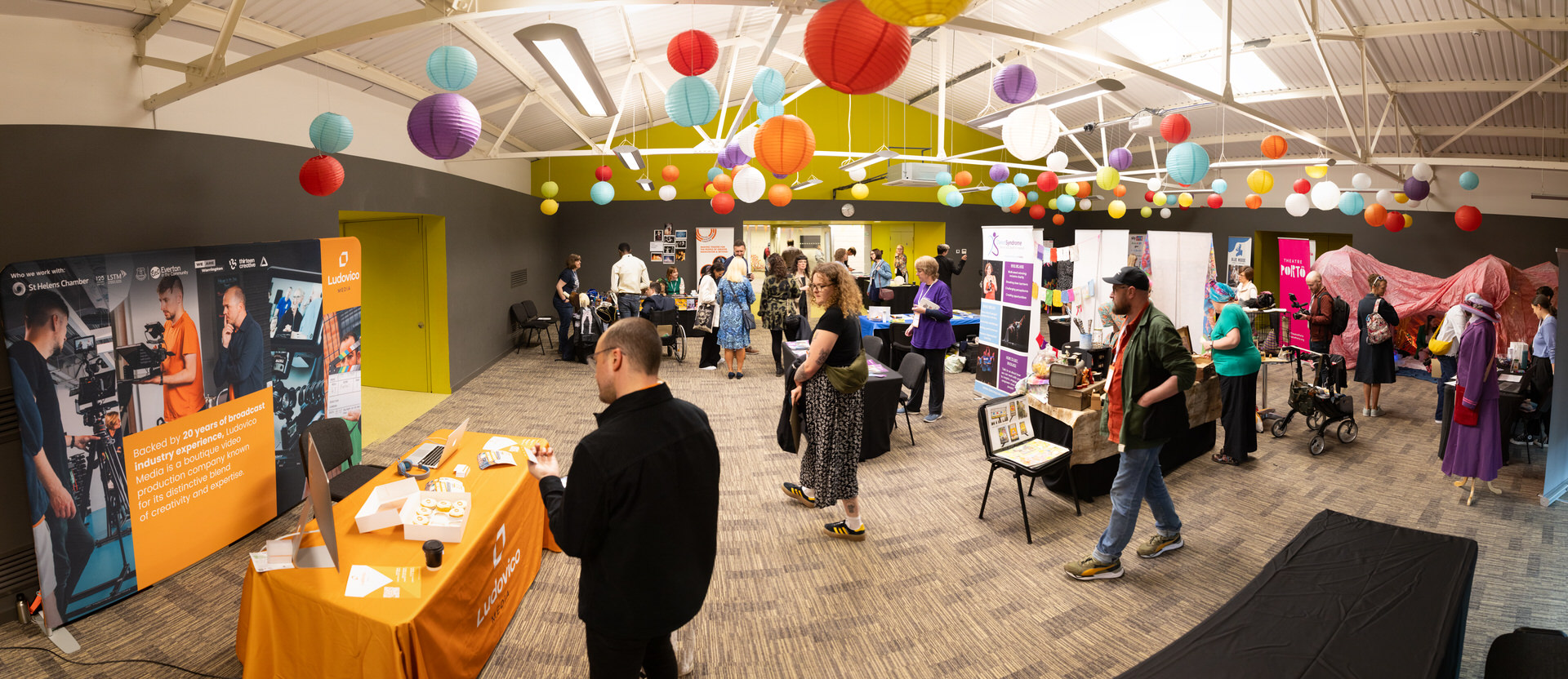 Image shows a colourful cultural marketplace. The cieling is decorated with multi-coloured paper lanterns and many people are milling around the stalls.
