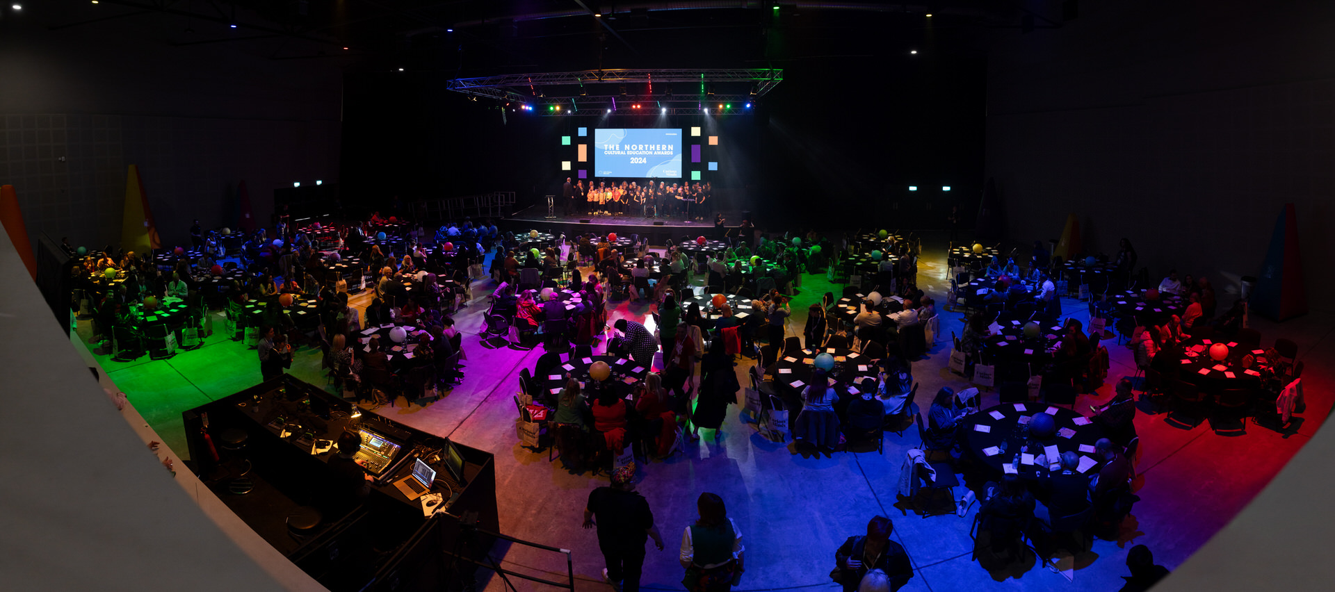 Image shows a large area, colourfully lit with cabaret-style seating. There is a performance happening on a stage in the background.