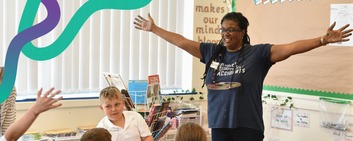 Image shows a Teaching Artist in a primary school classroom. The artist is in full flow, with arms outstretched, and a happy smile. The school pupils are engaged and enjoying their art lesson.
