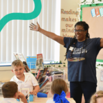 Image shows a Teaching Artist in a primary school classroom. The artist is in full flow, with arms outstretched, and a happy smile. The school pupils are engaged and enjoying their art lesson.