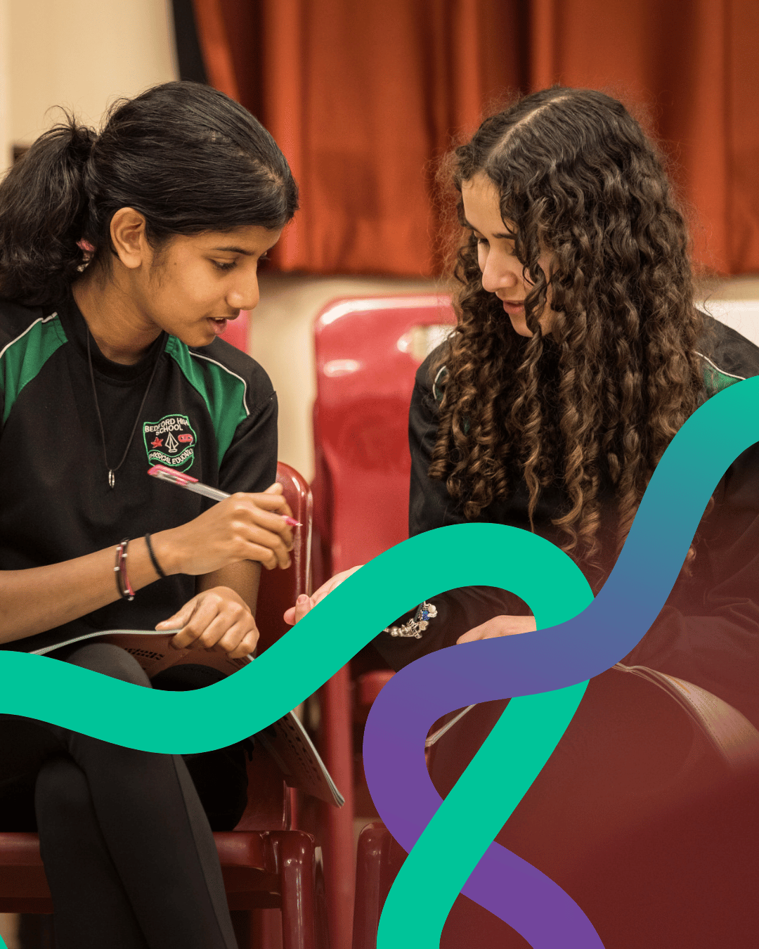 Image shows two secondary school pupils sitting next to each other in uniform. They are consulting a resource book and discussing their work